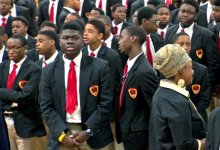 An assembly of teenage boys in tan slacks, white-collared, button-up shirts, black jackets, and red ties are standing. A female teacher is standing among them. 