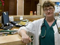 A female nurse is wearing green nursing scrubs, a white coat, and has a stethoscope around her neck. She's standing in a doctor's office behind a long counter next to her desk, and she's looking directly at the camera with a closed mouth smile.