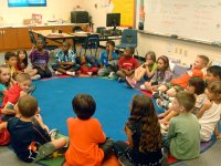 A group of young elementary students are sitting in an oval on their classroom floor on a blue circular rug with a colorful border.