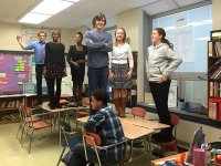 Six preteen students are standing on their classroom desks, smiling, and one student is sitting. 