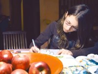 A young girl with glasses is sitting at her kitchen table writing. There's a bowl of apples on the table, next to an opened text book, next to an opened binder with lined paper.