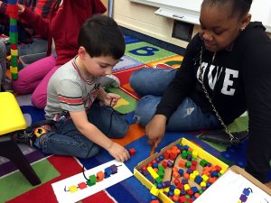 A fifth-grade girl is sitting on a colorful A, B, C carpet beside a young pre-school boy with special needs. The young boy is placing small, colorful, circular and square blocks with a hole in the center through a string.