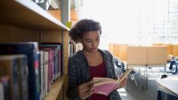 A young, teenage girl is leaning against a library shelf, reading a book.