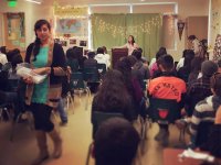 A high school classroom is decorated with white strands of lights, and a green sheet backdrop partially covering the front of the classroom. The classroom is filled with students sitting in blue chairs. One teacher is standing at a podium, addressing the 