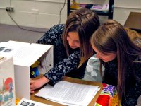 Two young women are squatting by a table in a classroom; one of them is reaching into a cardboard box that's part of a display.