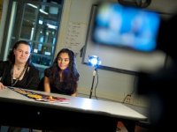 Two young girls are sitting at a table in a classroom being filmed with a light behind them.