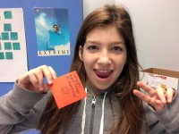 A young girl with long, brown-red hair is looking directly at the camera, smiling. She's standing in front of a classroom wall making a peace sign with her left hand and holding up an orange post-it in her right hand that says, "Computer aided design is a