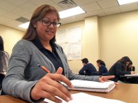 A closeup of a teenage girl sitting at her desk, smiling. One hand is resting on an opened notebook, and her other hand is holding a pencil above a sheet of paper she's writing on. Other students are sitting throughout the classroom.