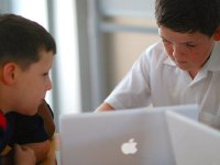 Two young boys are sitting next to each other in class with laptops in front of them.