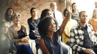 A group of adults sit and face someone outside the frame as one woman raises her hand to speak.