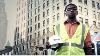 Boy wearing a yellow safety vest holding his hard hat, standing on a busy street in front of a very large building