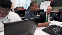 Man crouched down talking to a student who is sitting at his desk