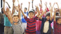Children in a classroom raise both hands in the air during a movement break.