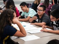 A group of young teenagers are sitting at their class table, working on a paper and pencil assignment, smiling, and talking.