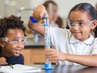 Two young students wearing safety goggles; one pouring liquid into a beaker