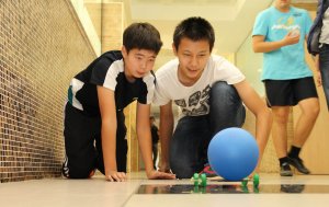 Two boys are kneeling in a school hallway, watching a blue ball roll on a paper-stick contraption with wheels.