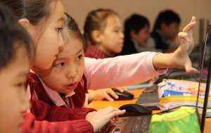 Two young girls are sitting, pointing and talking about something on a laptop screen. 