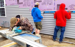 One boy at a table saw and two boys leveling and installing siding on the house