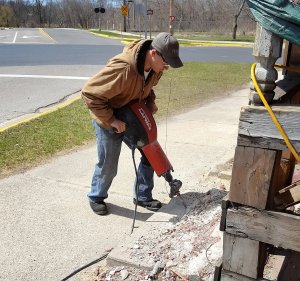 Boy wearing safety goggles and heavy boots using a jack hammer to break up concrete next to the house and sidewalk