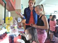 Teacher standing talking with a student as they watch a girl pouring liquid into cups at a counter