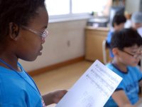 A closeup, side profile of a young girl with glasses standing in a classroom, reading a music sheet.