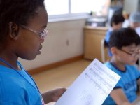 A closeup, side profile of a young girl with glasses standing in a classroom, reading a music sheet. 