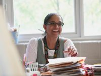 A female teacher is sitting at a desk, looking up and smiling. She's holding a pen in one hand, and stacks of paper and folders are in front of her. She's wearing glasses, big, pearl-like earrings, a turquoise necklace, and a lanyard.