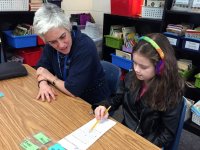 An young girl with rainbow-knitted headphones is sitting at a table with her female teacher. They're both looking at a long, thin sheet of paper, and the young girl is holding the tip of the pencil against the paper, pointing at something.