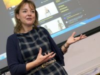 A closeup of a female teacher standing in front of a projector. It looks as if she's addressing her class, speaking, with her hands out, gesturing.