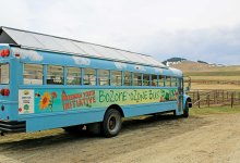An exterior shot of a blue-painted bus with clouds and a solar panel roof. It's parked on dirt overlooking hills.