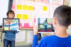 A young boy is using an iPad to take a photo of another young boy holding up his artwork.