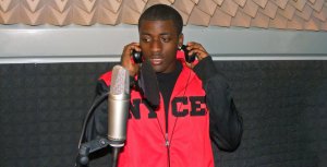 A teen male wearing headphones is standing next to and speaking into a microphone.