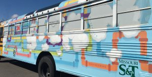 A close-up side exterior of a school bus painted blue with white clouds and purple, pink, green, yellow, and blue pipes.