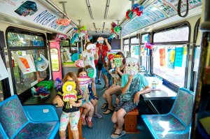 A group of kids and adults are sitting in a bus, holding Halloween masks they created.