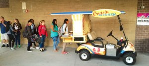 Students are lined up behind a golf cart filled with food that says, 'Cruisin' Kitchen.'