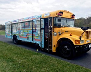 A side exterior shot of a school bus. The majority of the bus is painted blue with colorful piping and white clouds. 