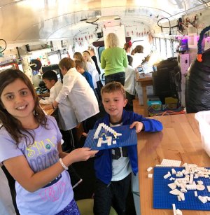 Elementary-aged students in lab coats are making things with iPads and Legos inside of a bus converted into a work station. 