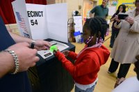 An elementary school girl submitting a mock election ballot in a school gym and receiving an "I Voted" sticker from a teacher
