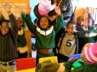 A group of eight young, elementary students are standing by their classroom desks with their arms raised in the air, talking. An adult is standing by them, holding a baby.