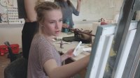 A female teen is sitting in an art class, holding a long paint brush, painting on a canvas.