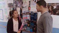 A female teen is standing in a school hallway talking to a male teacher; there are dance fliers on a bulletin board on the wall beside them.