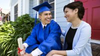 A teen boy in a graduation cap and gown sits on the front porch with his mom—both are smiling. 