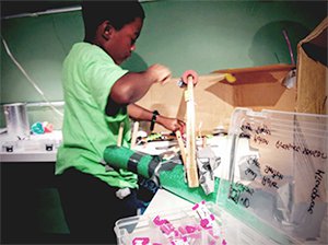 A young boy in a green t-shirt and black pants is at a work station covered with cardboard boxes and a plastic container filled with white and pink plastic building blocks. He's attaching two long, rectangular pieces of plastic, forming a bridge.