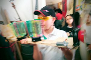 A young boy wearing a black and red cap, standing in a classroom, is holding a wind turbine made out of plywood, duct tape, and a fan, and he's balancing yellow, green, and pink, clear plastic cups on top of the wind turbine.