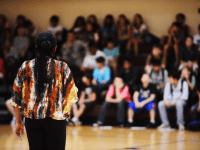 A photo of a woman talking to a large group of students in a school auditorium.