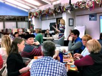 A group of adults are sitting among four tables, six to a table, in a library. Paper plates, popcorn bags, soda, and water bottles are on top of their tables, alongside open binders. A female adult is standing in front of everyone, addressing them.