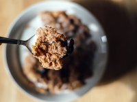 A closeup of a spoon with milk, corn flakes, and raisins. The rest of the image is out of focus. Beneath the spoon is a bowl filled with cereal on a light brown, wooden table. 