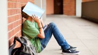 Middle school aged male student sitting with back to a wall holding an open book over his head and face. 