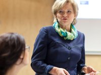 A short-haired blonde woman in her early sixties, wearing a blue coat and a blue, green, and yellow scarf, is standing in front of a projector, talking to a seated brunette woman wearing glasses.