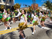 A street parade is going on in a downtown area. A lot of girls in white shirts and green shorts are walking in the street with their hands raised, carrying golden pom-poms.